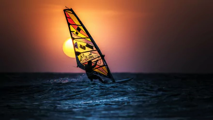 HD desktop wallpaper showing windsurfing on the ocean at sunset, with vibrant orange sky and sun near the horizon over the sea.