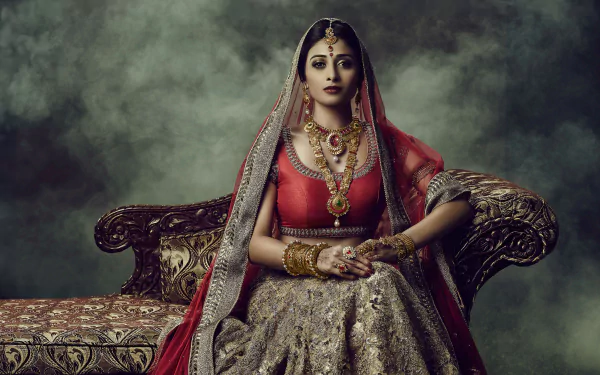 A high-definition desktop wallpaper featuring an Indian bride adorned in a saree, necklace, bracelet, and intricate jewelry, sitting on a traditional ornate sofa with a misty background.