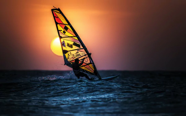 HD desktop wallpaper showing windsurfing on the ocean at sunset, with vibrant orange sky and sun near the horizon over the sea.