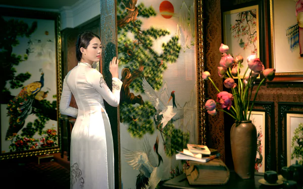 A Vietnamese woman in a white Ao Dai stands beside a painted screen with cranes, holding a book, surrounded by floral paintings and a bouquet in a vase, captured in 4K Ultra HD.