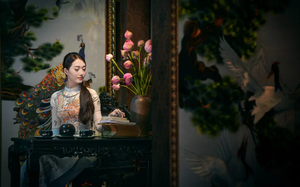 A Vietnamese woman in an Ao Dai sits at a table with a tea set, book, and necklace, surrounded by flowers and a traditional painting, captured in 4K Ultra HD.