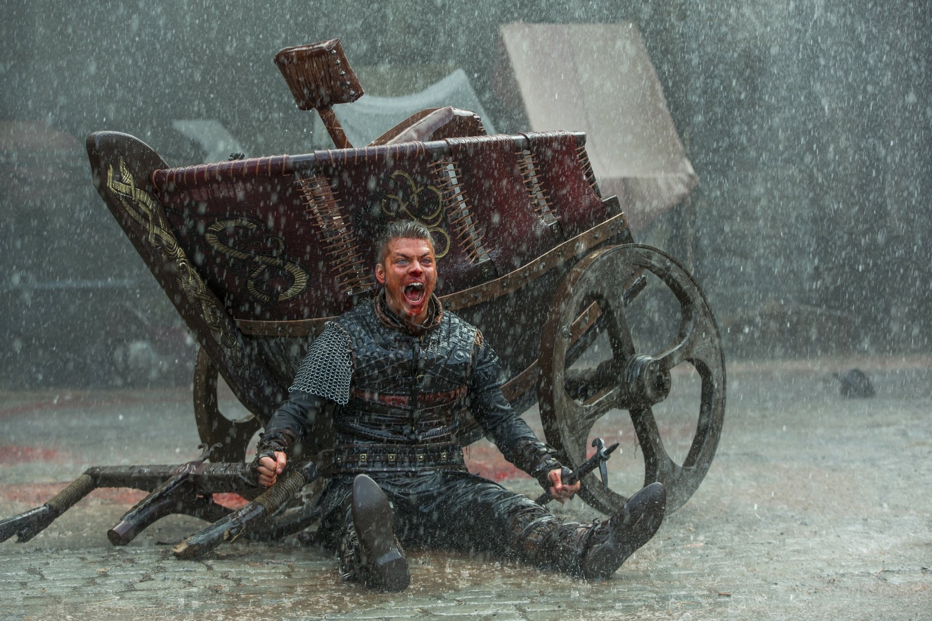 A fierce scene from Vikings (2013) TV show in 4K Ultra HD, depicting a warrior seated on the ground in the rain beside a broken chariot, expressing intense emotion.
