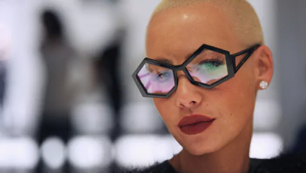 A close-up HD wallpaper of a woman with bold hexagon-shaped glasses, shaved head, and red lips, exuding a confident persona.