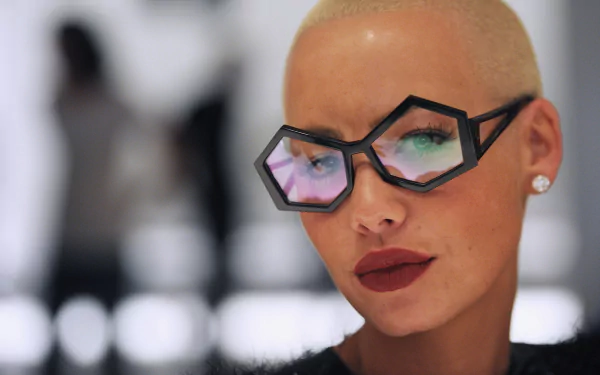 A close-up HD wallpaper of a woman with bold hexagon-shaped glasses, shaved head, and red lips, exuding a confident persona.