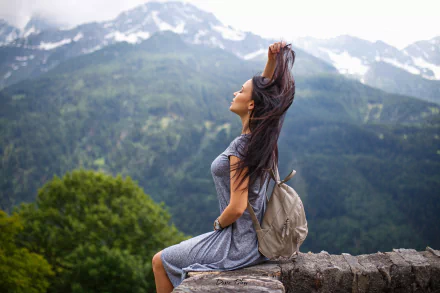 HD desktop wallpaper featuring a woman model sitting on a stone ledge with mountains and greenery in the background, hair lifted by the breeze.