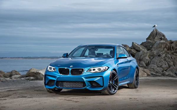 Blue BMW M2 car parked on a rocky beach under a cloudy sky, captured in 4K Ultra HD as a PC desktop wallpaper and background.