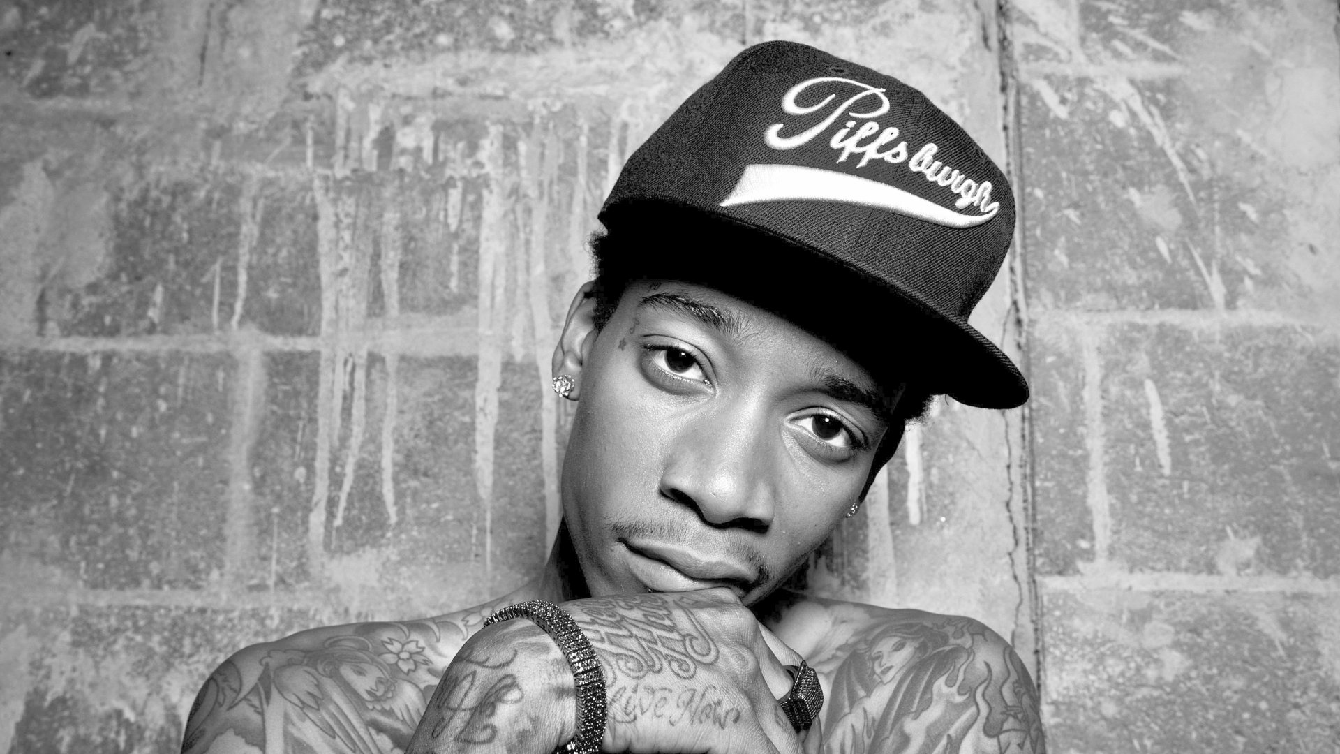 HD desktop wallpaper featuring a monochrome portrait of a man with tattoos wearing a Pittsburgh cap against a brick wall background.