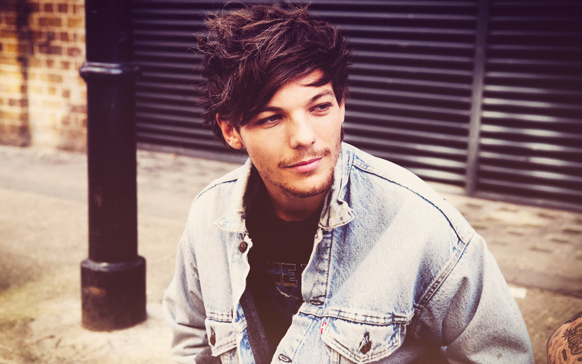 HD desktop wallpaper featuring Louis Tomlinson, sitting casually with a stylish denim jacket, against an urban backdrop, conveying a laid-back vibe in a music-inspired setting.