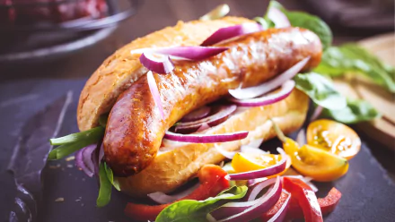 A close-up HD desktop wallpaper of a hot dog with a grilled sausage, fresh greens, sliced red onions, and cherry tomatoes in a bun.