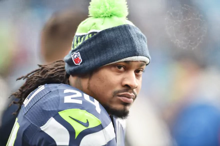 Marshawn Lynch, former Seattle Seahawks star, gazes intently while wearing his team gear and a pom-pom beanie, capturing the essence of sports commitment in this HD wallpaper.
