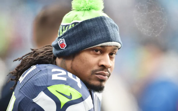 Marshawn Lynch, former Seattle Seahawks star, gazes intently while wearing his team gear and a pom-pom beanie, capturing the essence of sports commitment in this HD wallpaper.