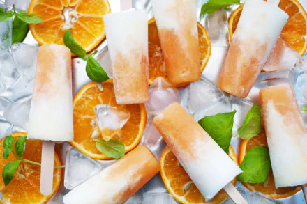 4K Ultra HD wallpaper featuring orange and cream ice cream popsicles surrounded by ice cubes and fresh orange slices, highlighting vibrant fruit and sweet treats.