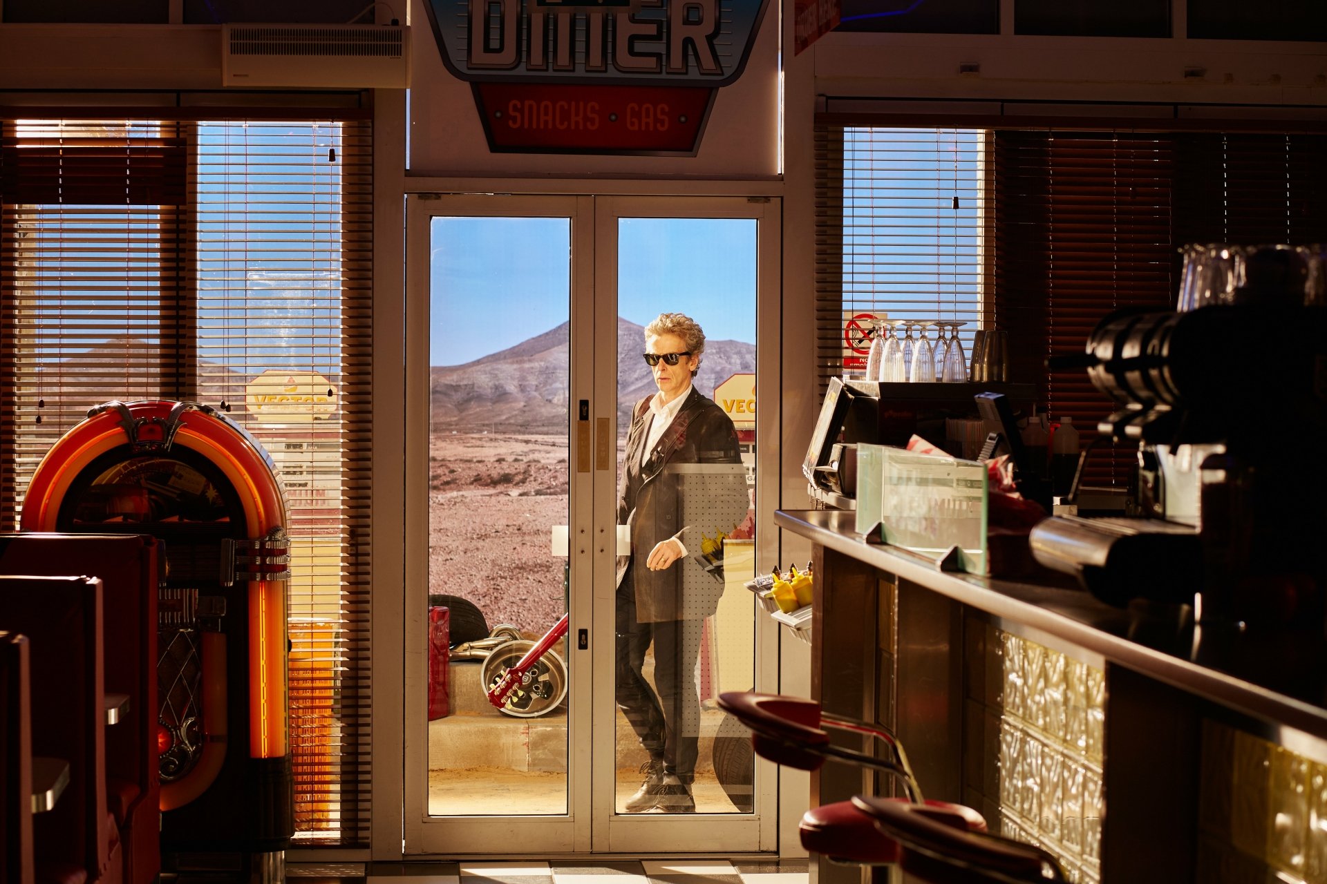 Peter Capaldi as the 12th Doctor stands outside a diner in a desert setting, featured in a 4K Ultra HD Doctor Who (2005) TV show desktop wallpaper.