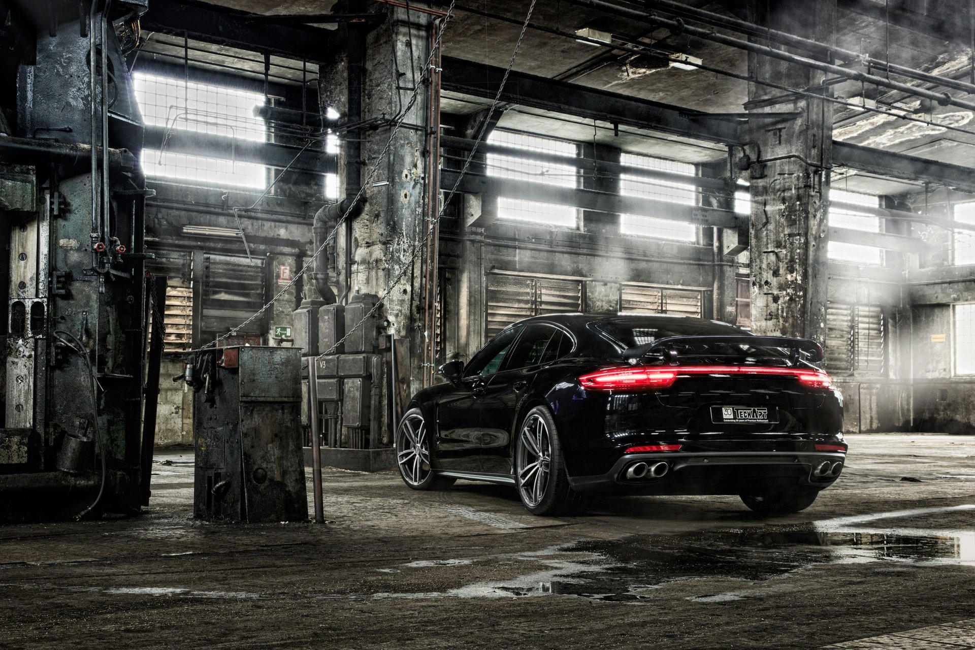 A black Porsche Panamera parked inside an abandoned factory. The image is a HD desktop wallpaper showing the vehicle in a gritty, industrial setting, with beams of light filtering through broken windows.