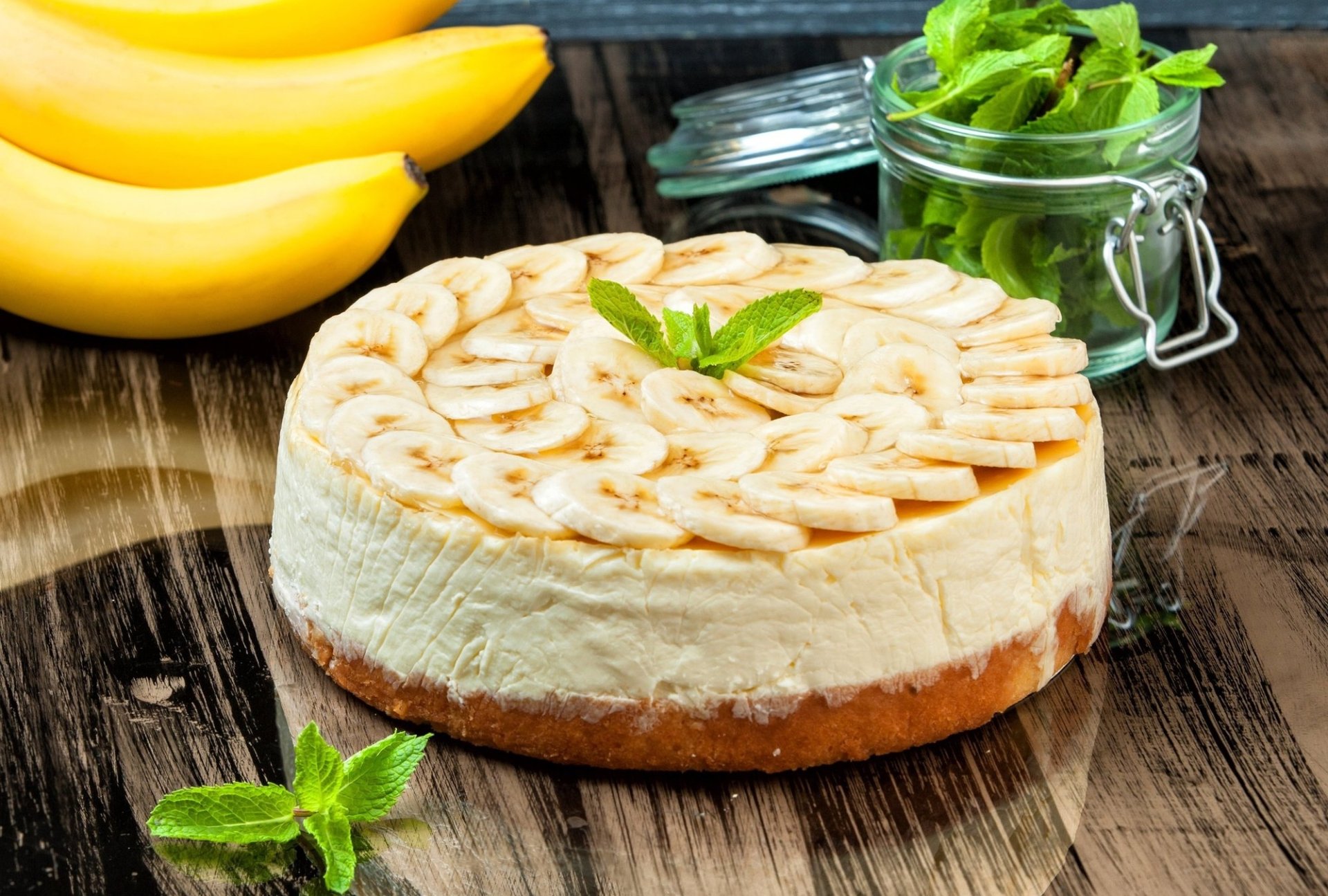 HD desktop wallpaper showcasing a banana pastry cake garnished with fresh mint on a wooden surface, accompanied by whole bananas and a jar of mint leaves.