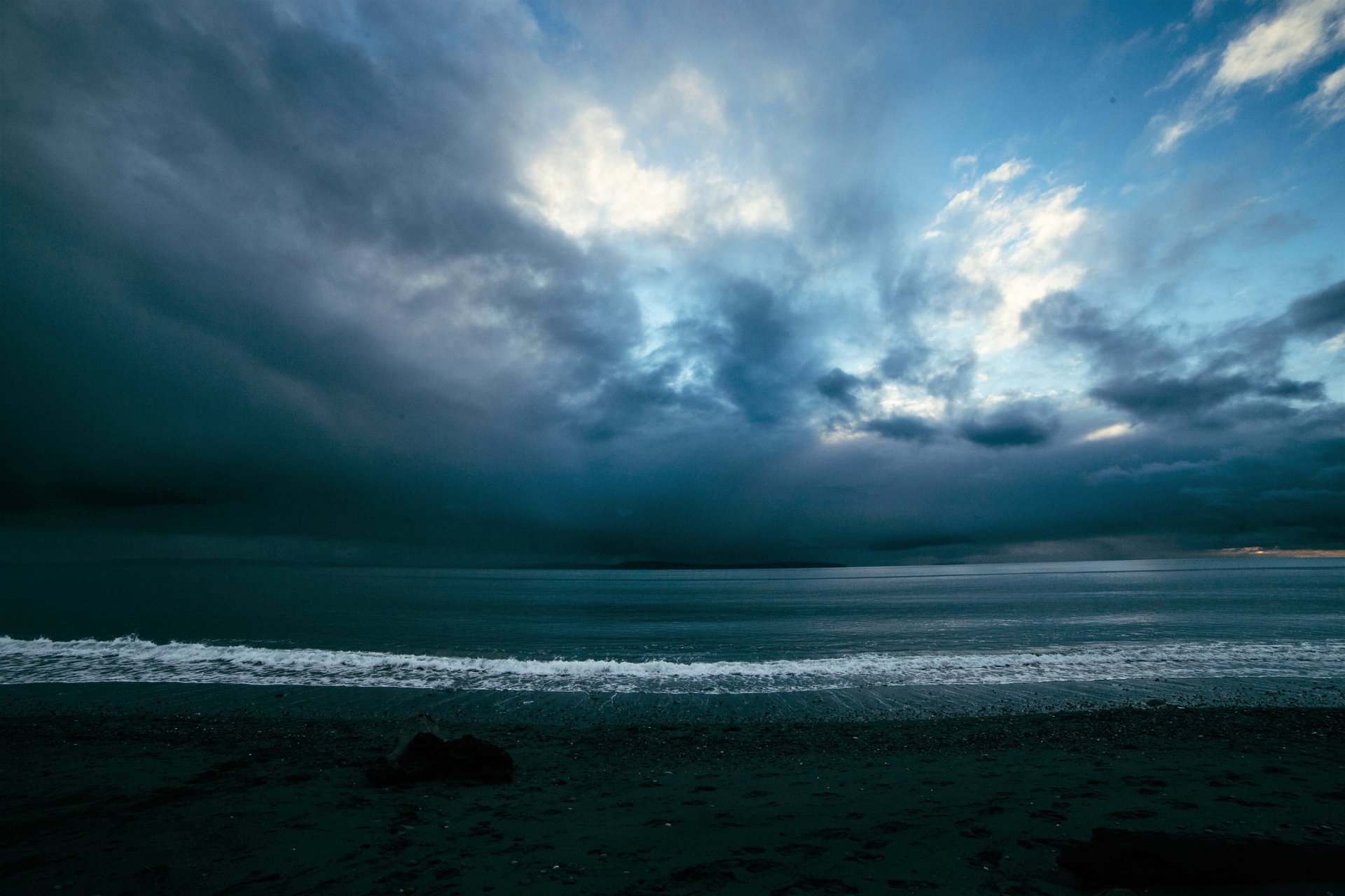 A dramatic seascape featuring a stormy ocean under a blue-gray sky, with swirling clouds and gentle waves lapping at the shore, creating a striking natural backdrop.