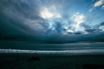 A dramatic seascape featuring a stormy ocean under a blue-gray sky, with swirling clouds and gentle waves lapping at the shore, creating a striking natural backdrop.