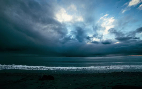 A dramatic seascape featuring a stormy ocean under a blue-gray sky, with swirling clouds and gentle waves lapping at the shore, creating a striking natural backdrop.