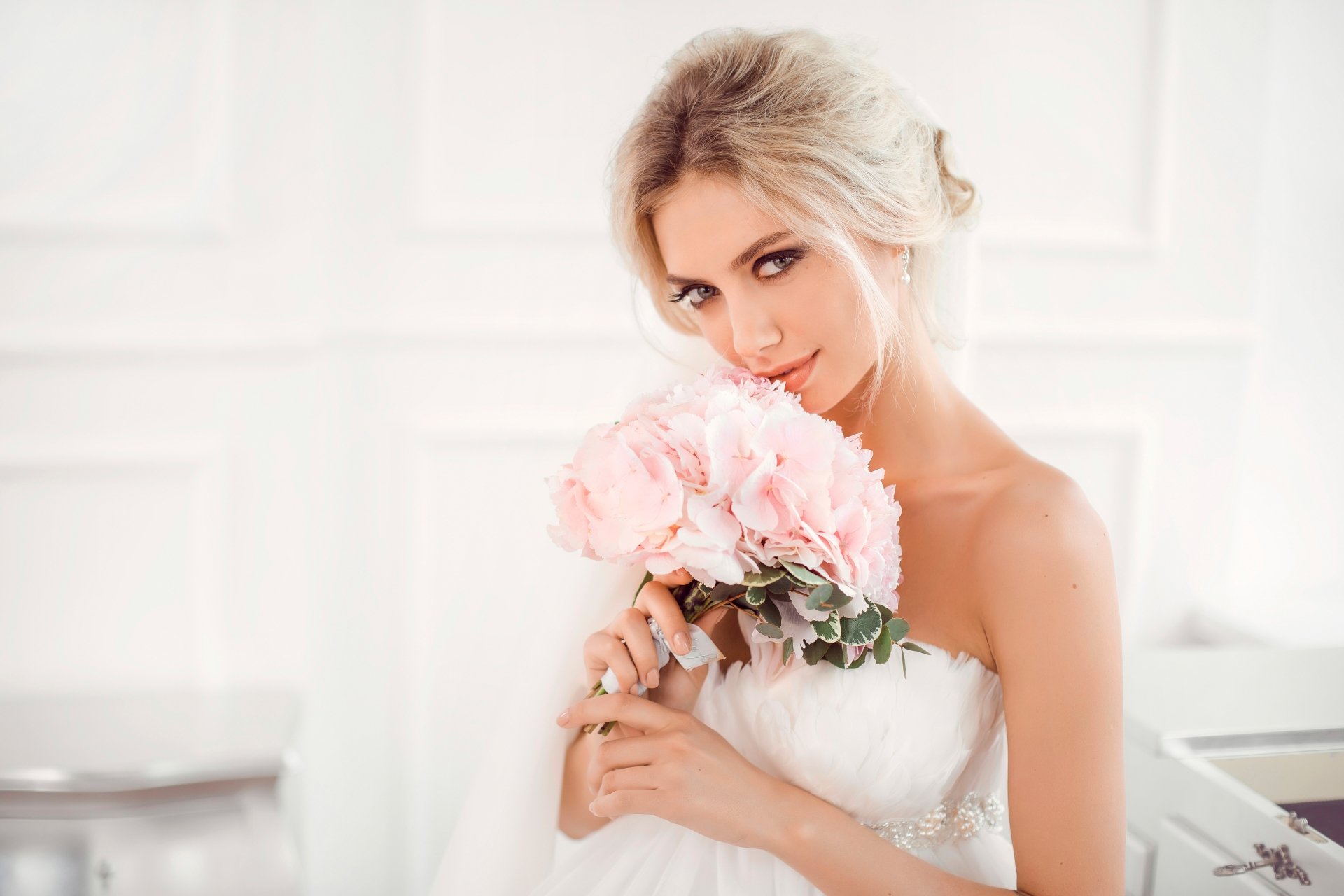Blonde bride with blue eyes gently holding a pink bouquet, captured in a soft, elegant 4K Ultra HD wallpaper.