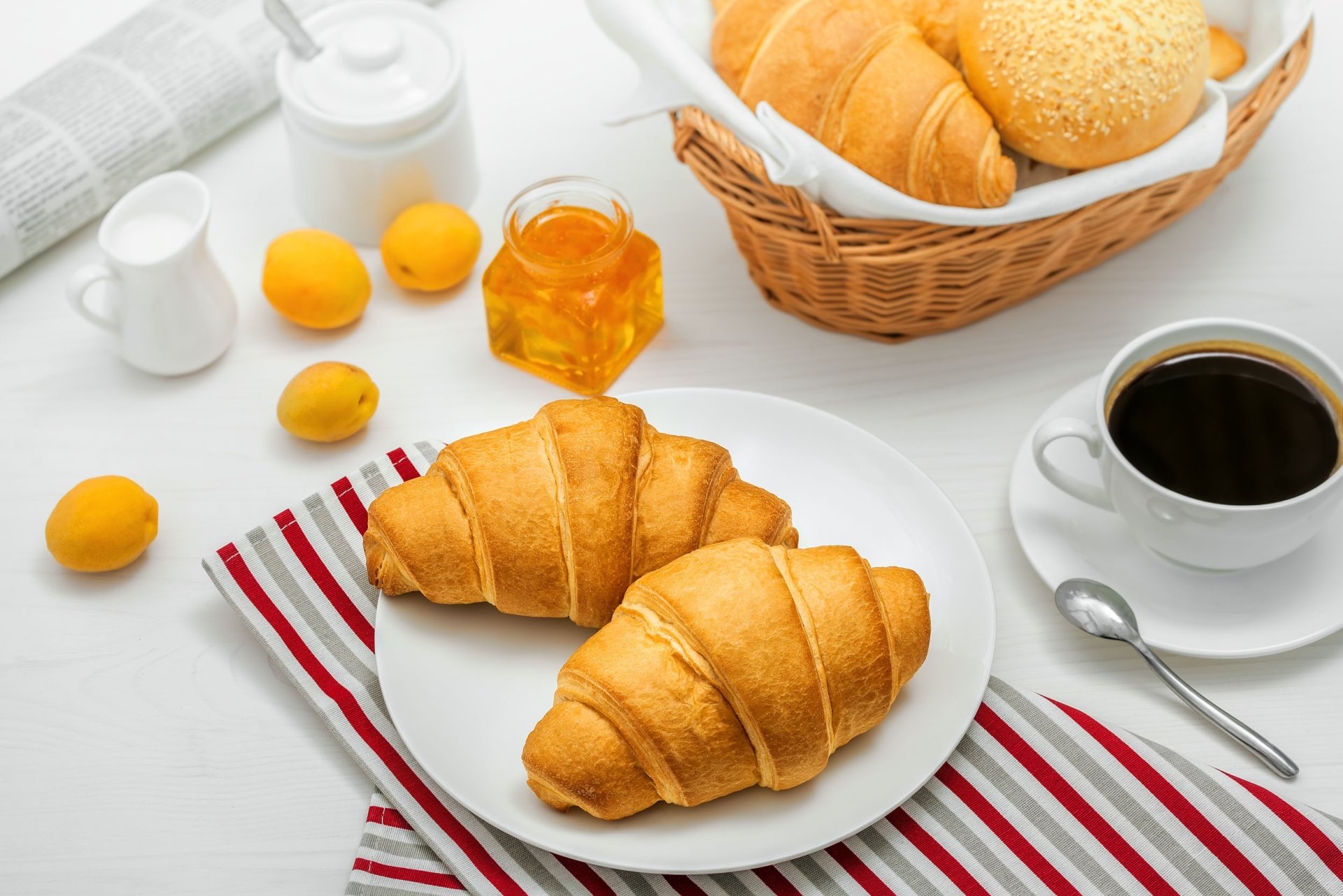 HD PC desktop wallpaper: breakfast spread with two golden croissants, viennoiserie basket, lemons, milk jug, honey jar, and a cup of black coffee.