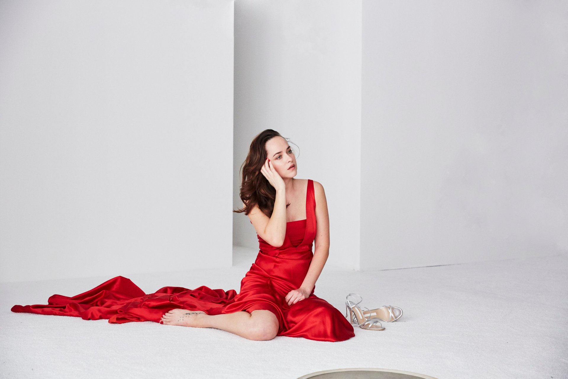 Brunette American actress Dakota Johnson in a red dress, seated gracefully on the floor against a minimalist white background, HD desktop wallpaper.
