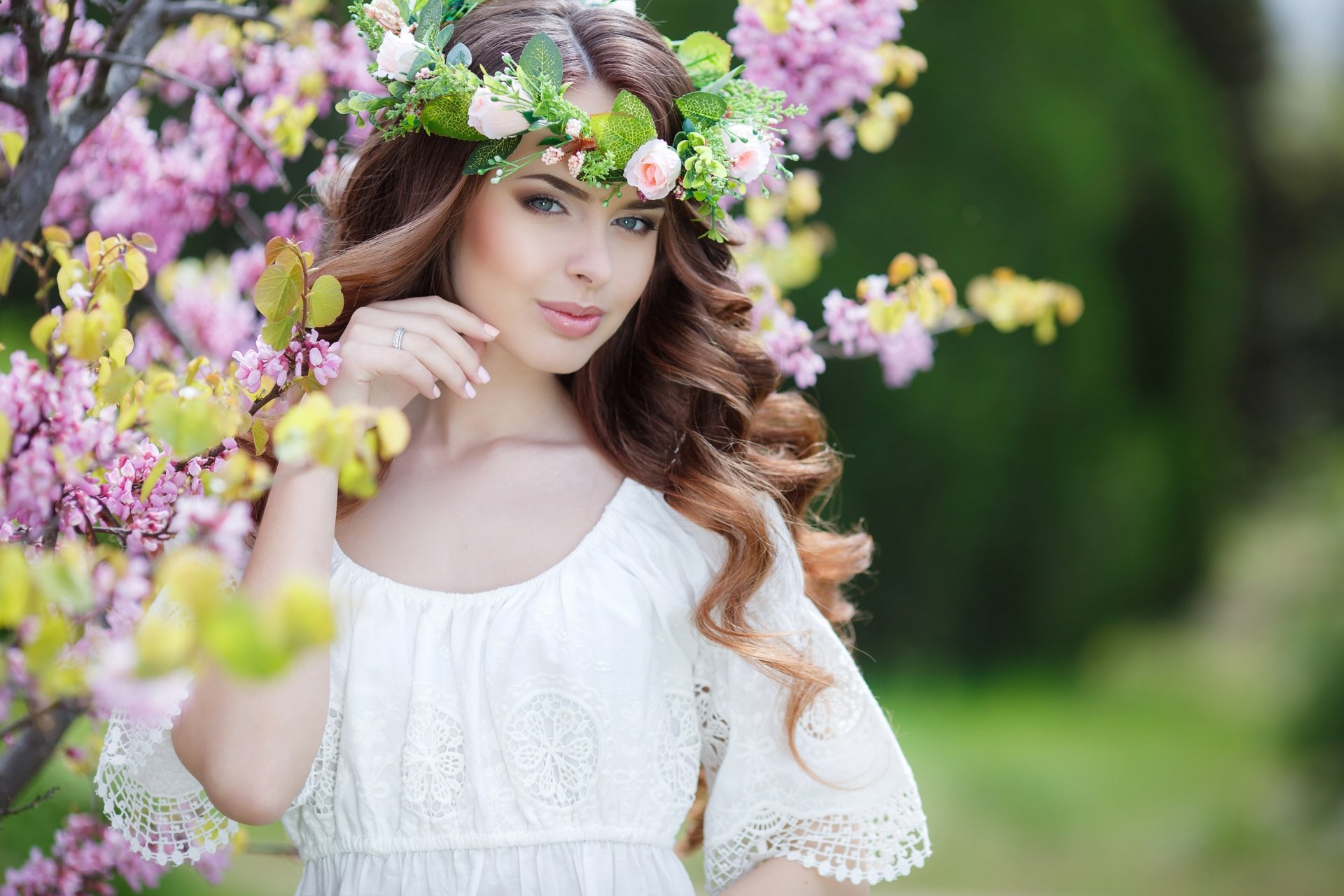 A brunette woman in a white dress and floral wreath poses among blossoming trees, captured with a shallow depth of field. This HD desktop wallpaper showcases the model in a serene, nature-filled setting.
