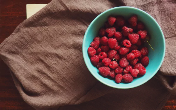 HD wallpaper of a bowl filled with fresh raspberries atop a rustic table with a linen cloth.