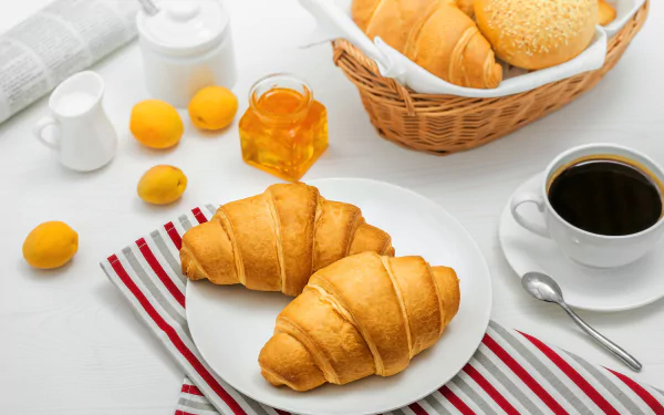 HD PC desktop wallpaper: breakfast spread with two golden croissants, viennoiserie basket, lemons, milk jug, honey jar, and a cup of black coffee.