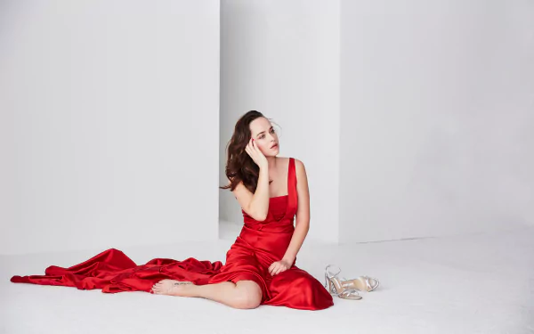 Brunette American actress Dakota Johnson in a red dress, seated gracefully on the floor against a minimalist white background, HD desktop wallpaper.