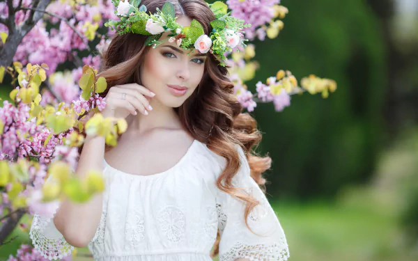 A brunette woman in a white dress and floral wreath poses among blossoming trees, captured with a shallow depth of field. This HD desktop wallpaper showcases the model in a serene, nature-filled setting.