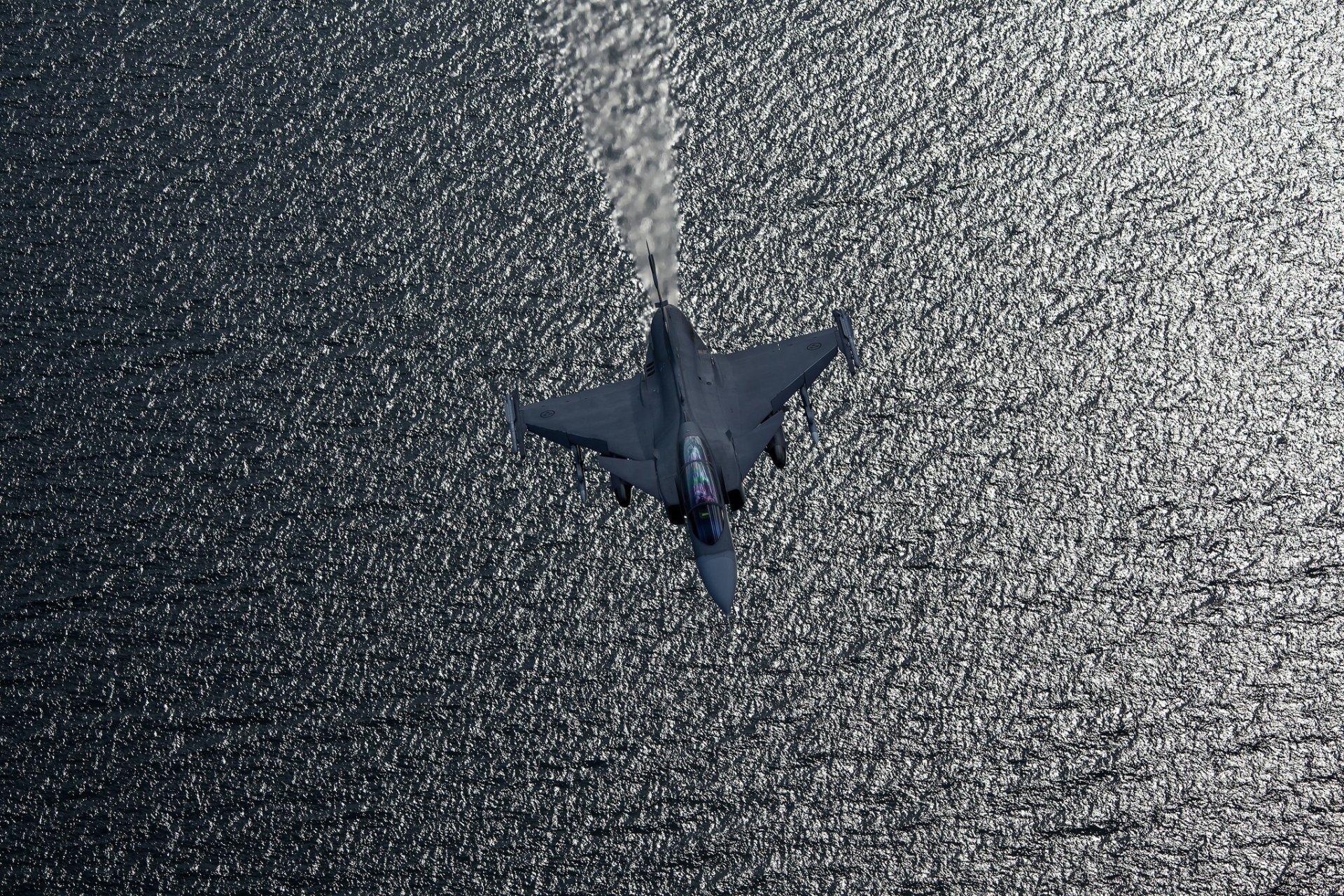A Saab JAS 39 Gripen jet fighter warplane flies over textured water, captured in a high-definition military aircraft desktop wallpaper.