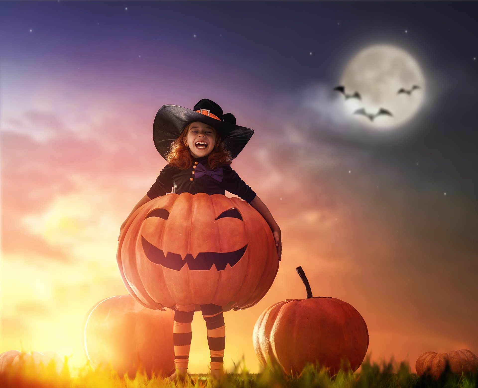 HD PC desktop wallpaper background: Halloween holiday scene with a smiling child in a witch hat inside a giant jack-o'-lantern, full moon and bats overhead.