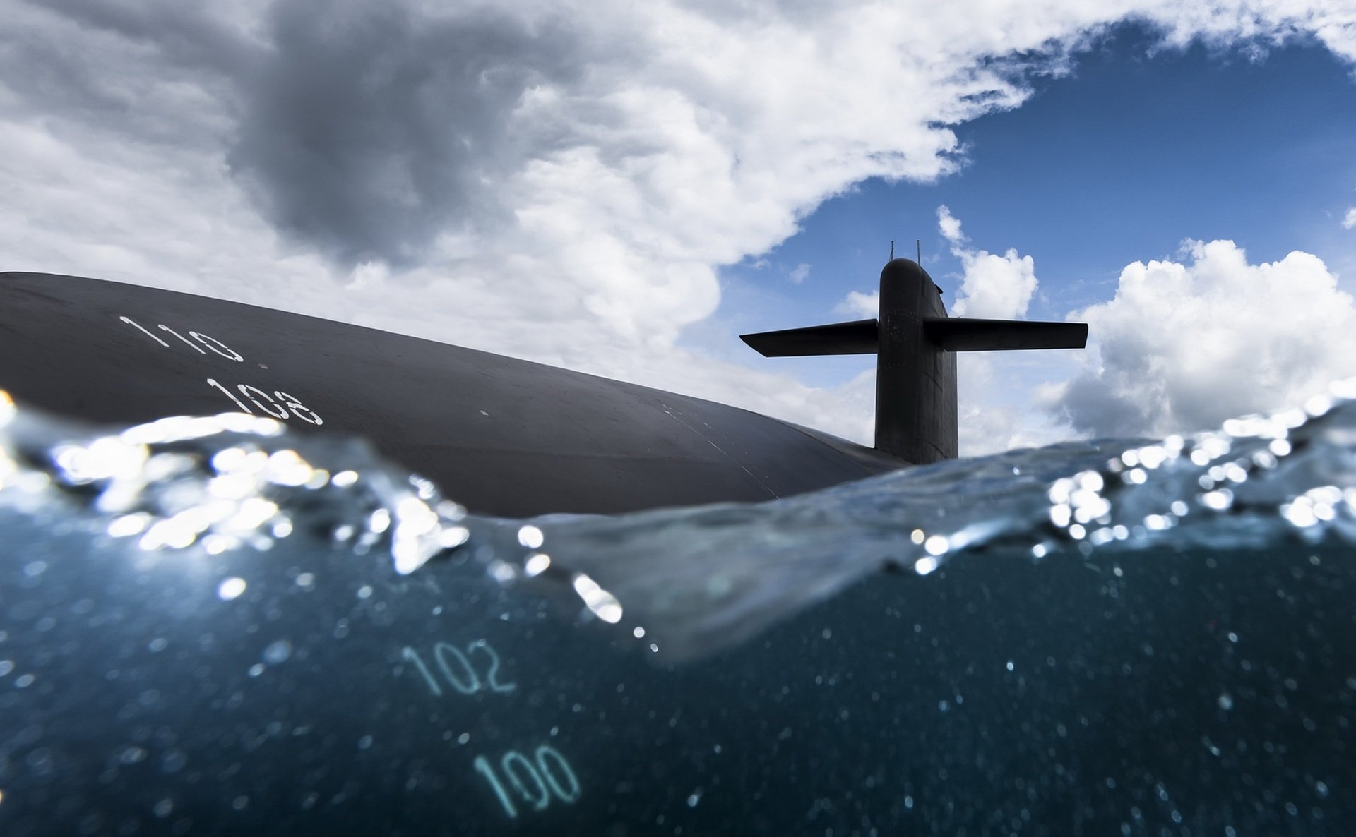 HD PC desktop wallpaper of a military submarine surfacing: sail and deck breaking choppy sea, depth markings visible through clear blue water beneath dramatic clouds.