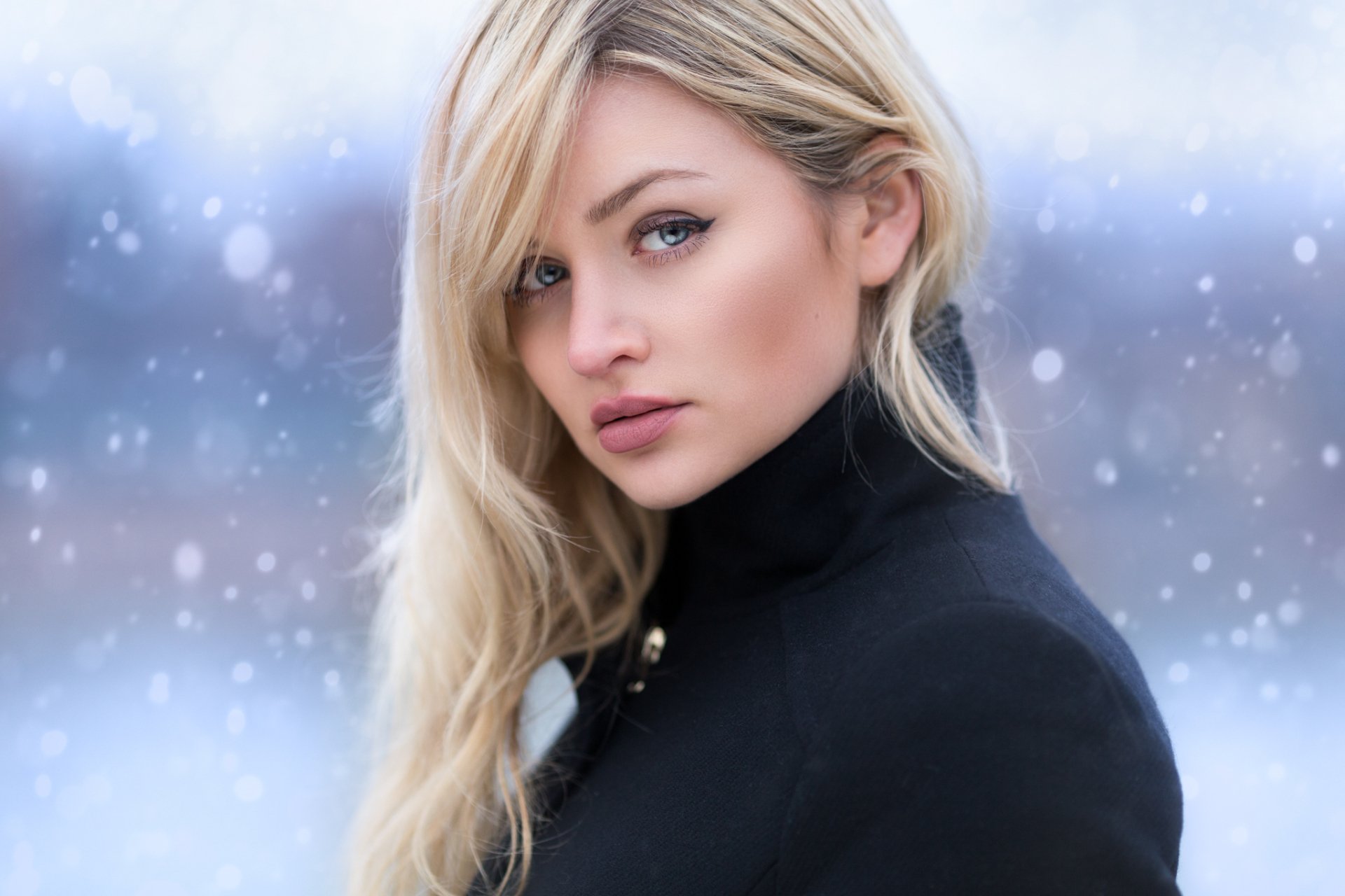 A blonde woman with blue eyes stands in a winter snowfall, wearing a black jacket. This HD desktop wallpaper captures the serene beauty of the model against a cold, snowy backdrop.
