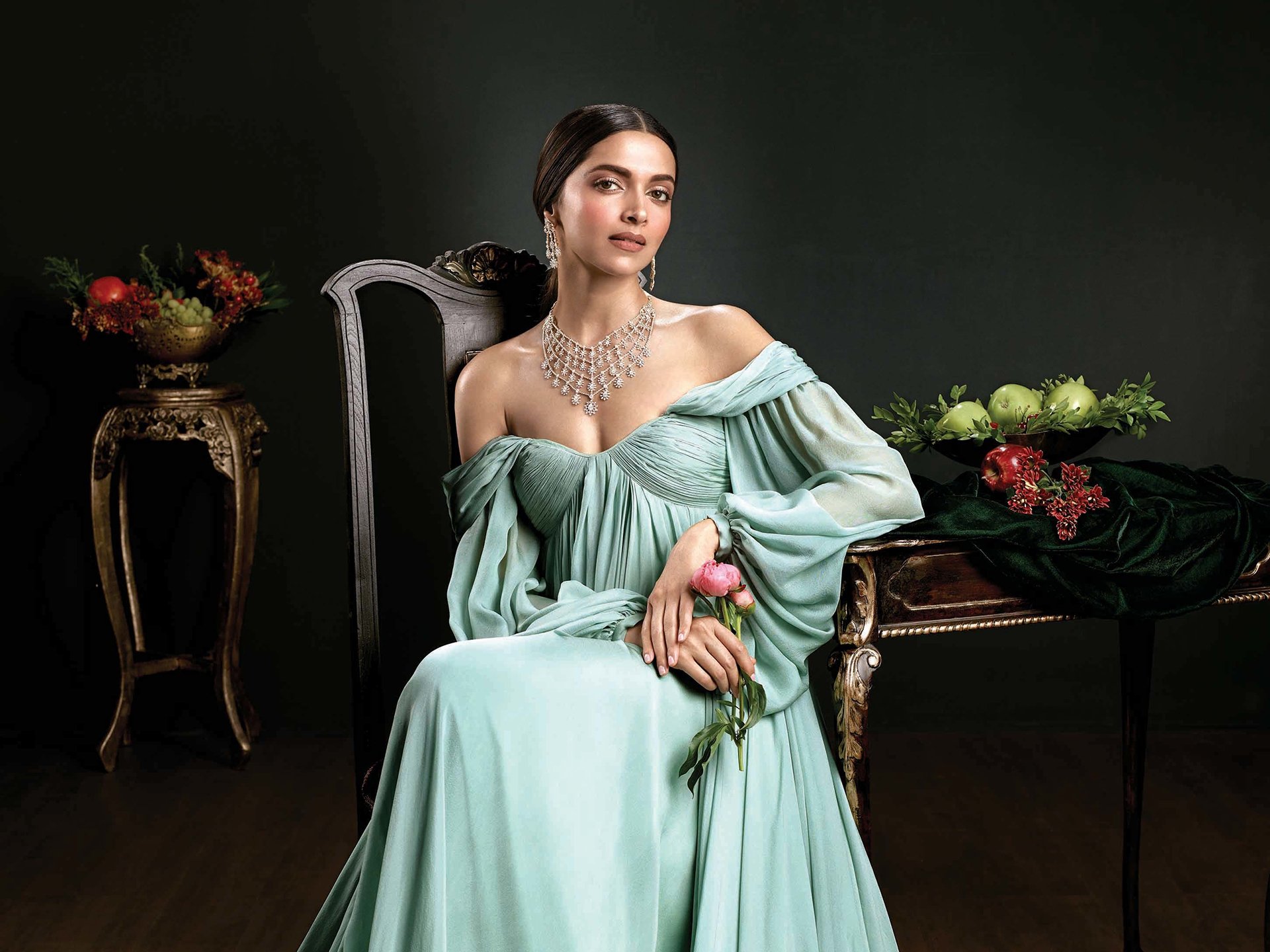 HD desktop wallpaper featuring celebrity Deepika Padukone in an elegant, off-shoulder pastel gown, sitting gracefully with floral and antique decor in a dark, moody setting.