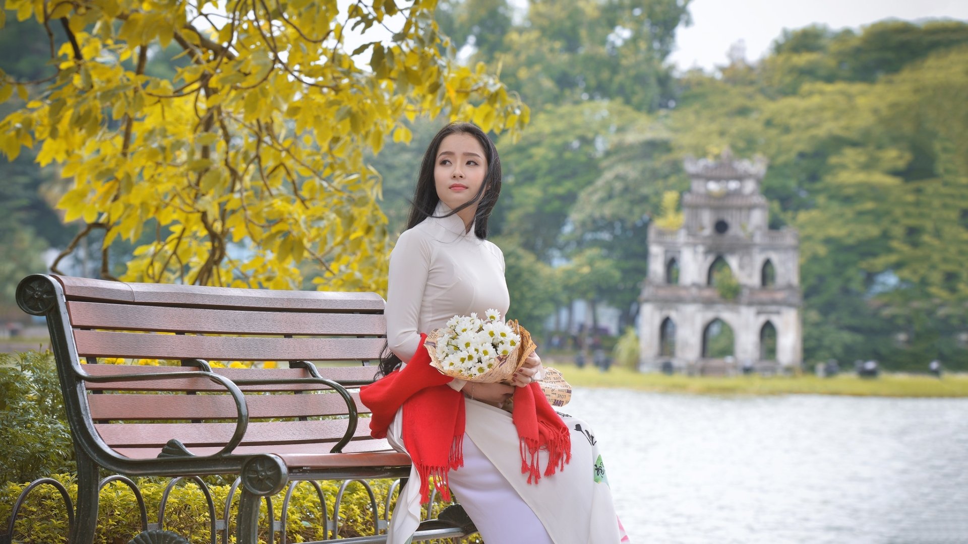 A brunette Asian woman in traditional costume sits on a bench by the water, holding a bouquet with a blurred historical structure in the background.