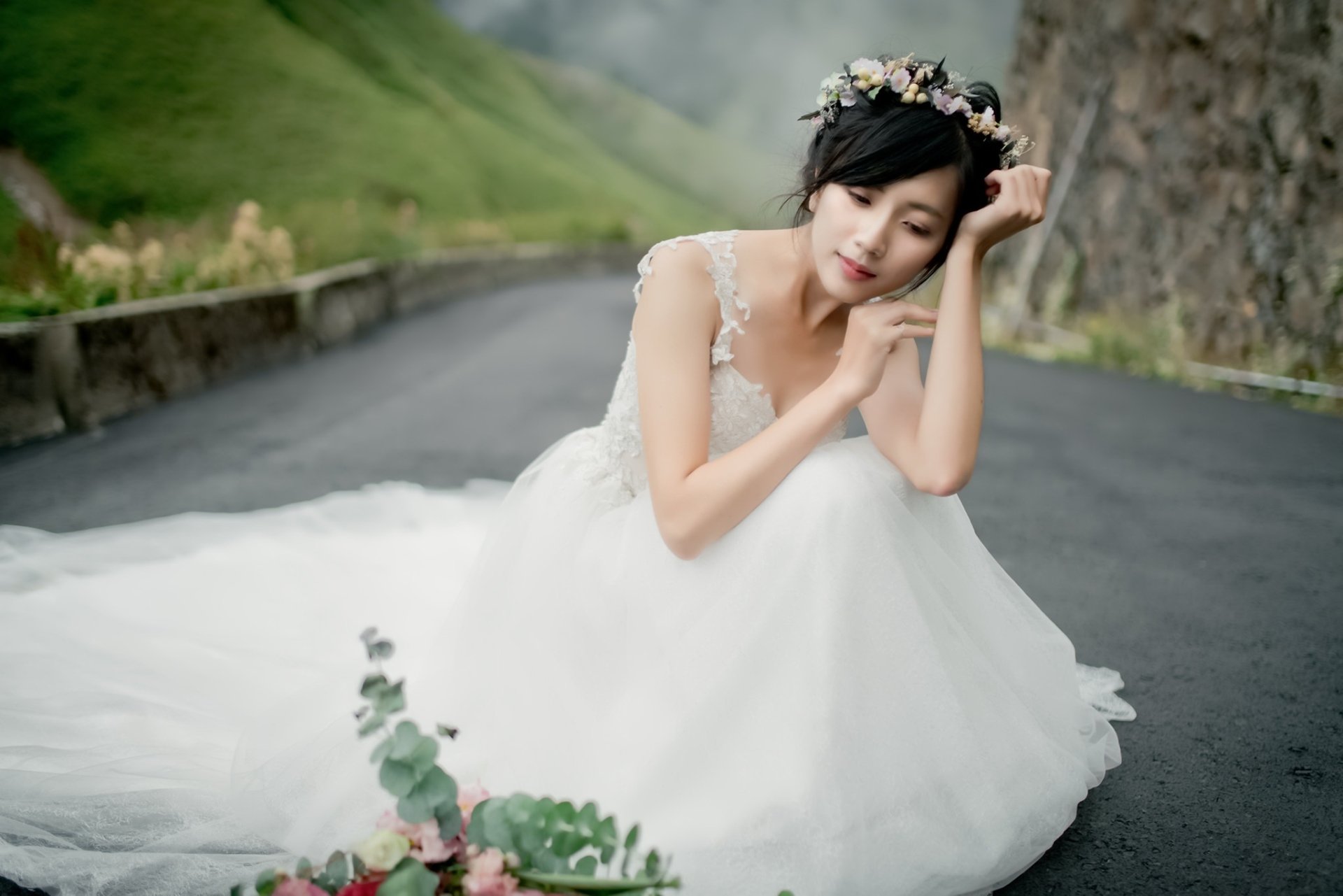 HD PC desktop wallpaper: bride, woman and model with short brunette hair and floral wreath, wearing a white wedding dress, seated on a road; soft background with shallow depth of field.