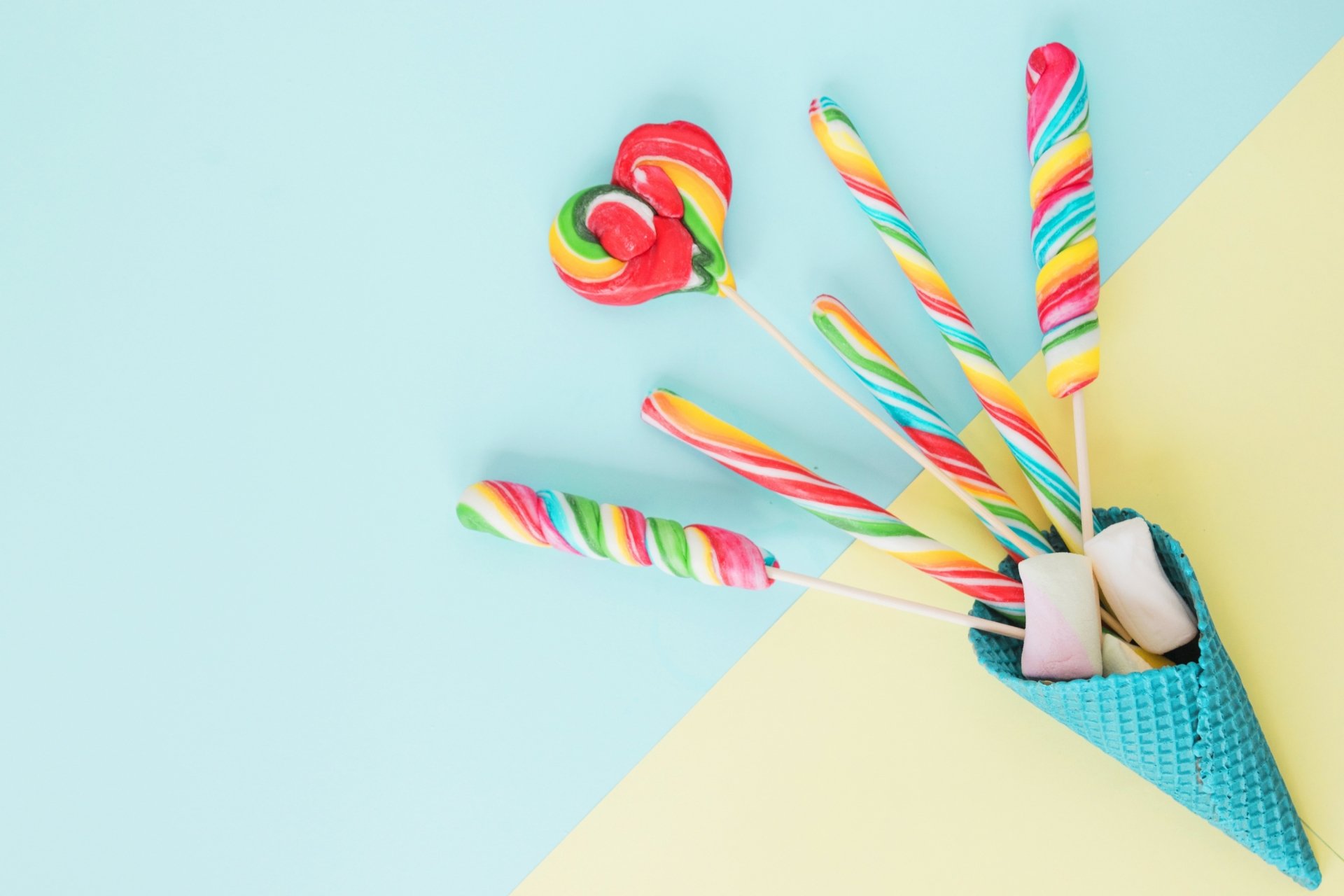 HD desktop wallpaper featuring colorful rainbow lollipops and sweets arranged in a blue waffle cone against a pastel blue and yellow background.