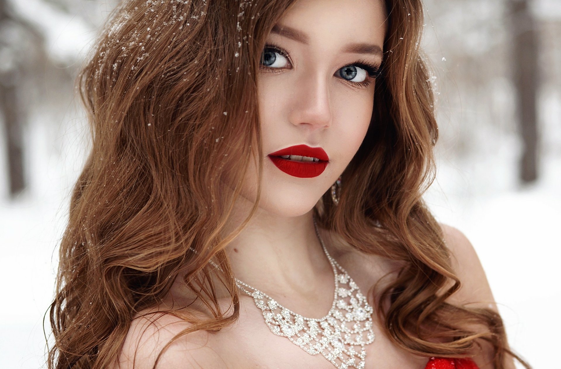 HD desktop wallpaper featuring a brunette woman with blue eyes, red lipstick, and a diamond necklace. She is standing in a snowy winter setting with snowflakes gently resting in her hair.