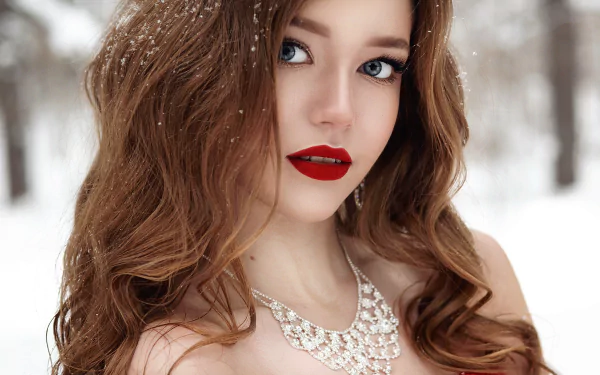 HD desktop wallpaper featuring a brunette woman with blue eyes, red lipstick, and a diamond necklace. She is standing in a snowy winter setting with snowflakes gently resting in her hair.