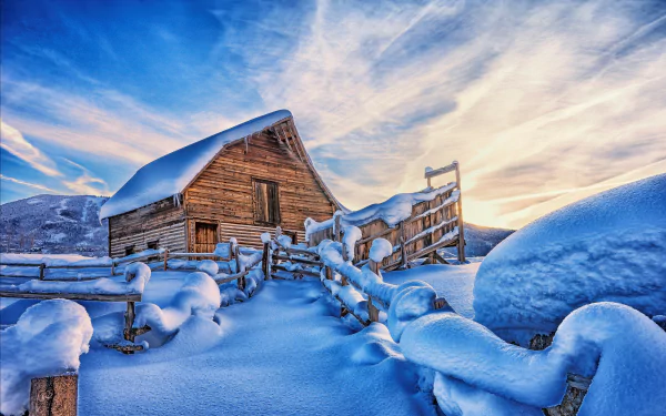 HD desktop wallpaper of a wooden cabin nestled in a snowy winter landscape with mountains in the background. The scene captures the serene beauty of snow-covered surroundings under a vibrant, partly cloudy sky.