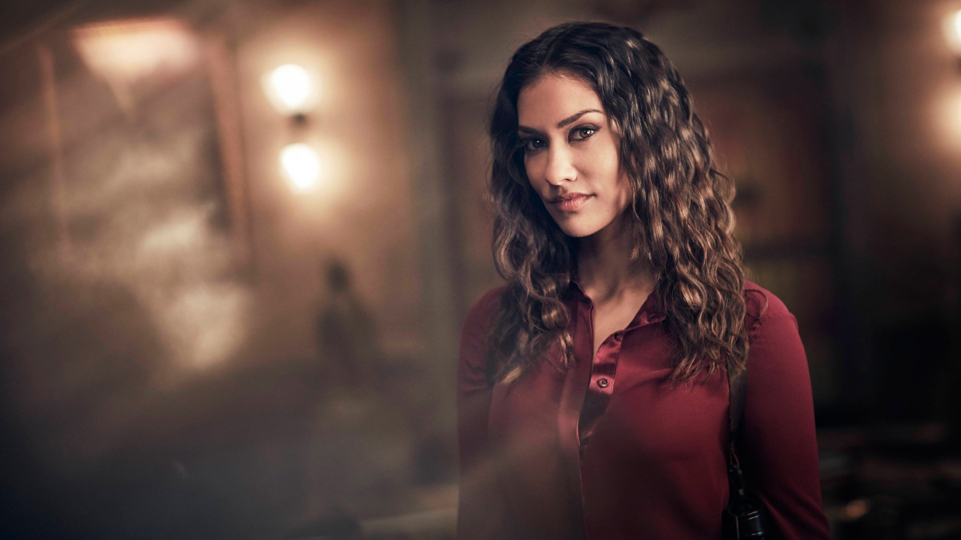 Portrait of Janina Gavankar with wavy hair, wearing a burgundy top, captured in a softly lit setting for a 4K Ultra HD PC desktop wallpaper.