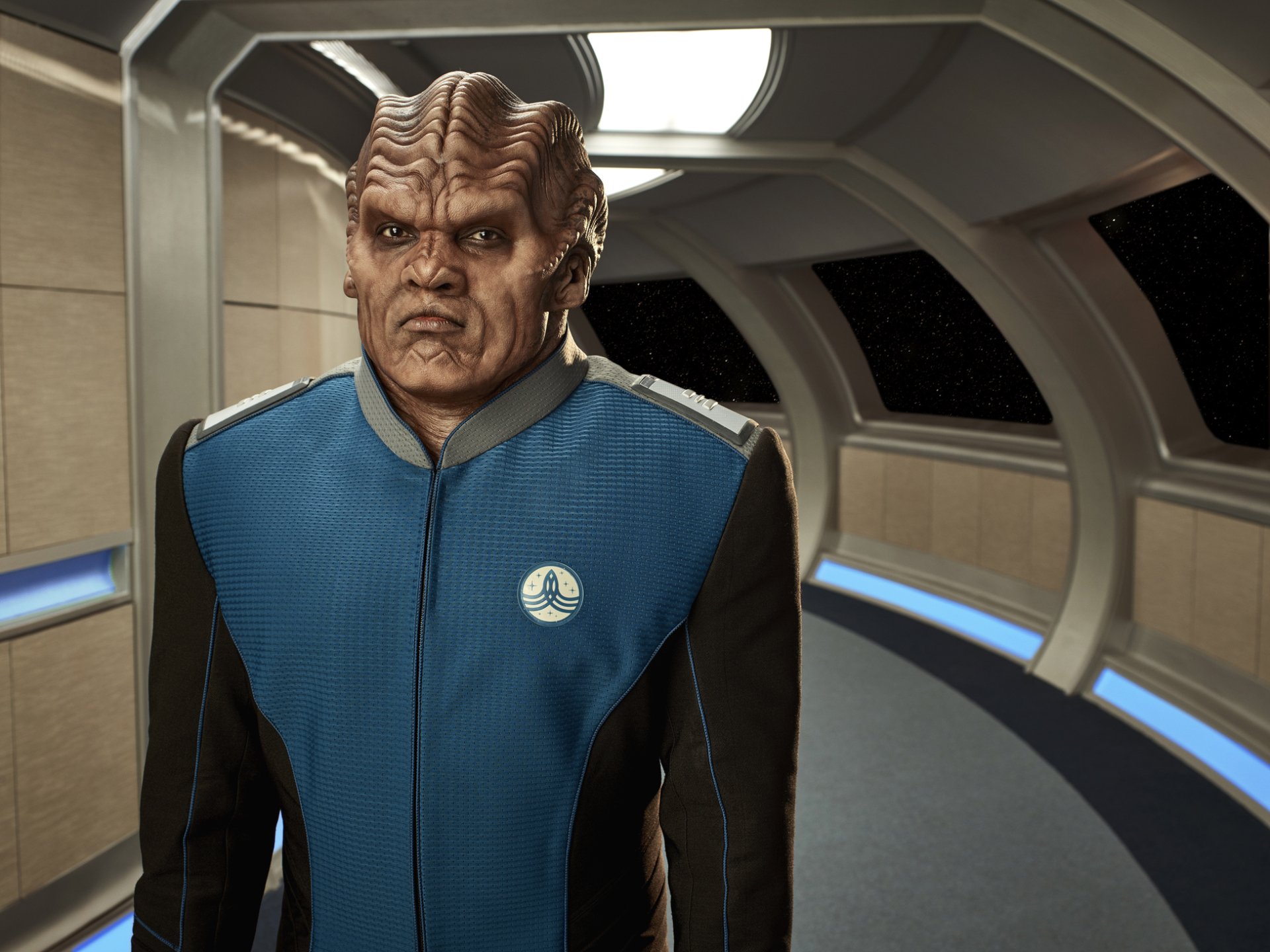 A character from The Orville stands in a sleek, starship corridor, showcasing intricate alien features and a blue uniform, set against a cosmic background. HD desktop wallpaper.