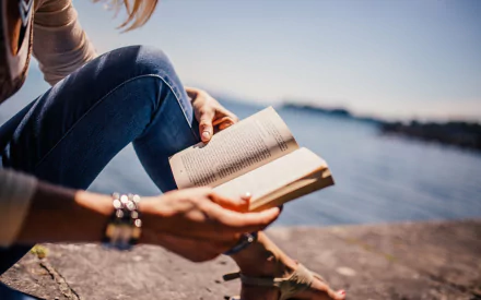 A person reading a book while sitting by the water, surrounded by a serene landscape. This HD desktop wallpaper captures a peaceful moment of leisure and reflection.