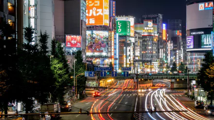 A vibrant night scene in Shinjuku, Tokyo, showcasing bustling streets adorned with bright advertisements and dynamic light trails from cars, set against a backdrop of towering buildings.