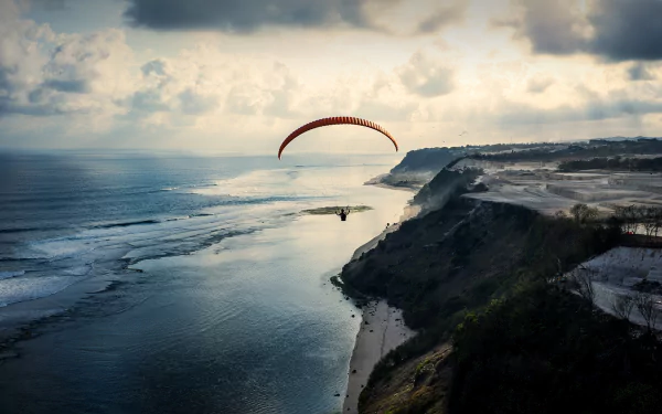 HD desktop wallpaper of a paraglider soaring over a beach coast with the ocean and horizon stretching under a dramatic cloudy sky.