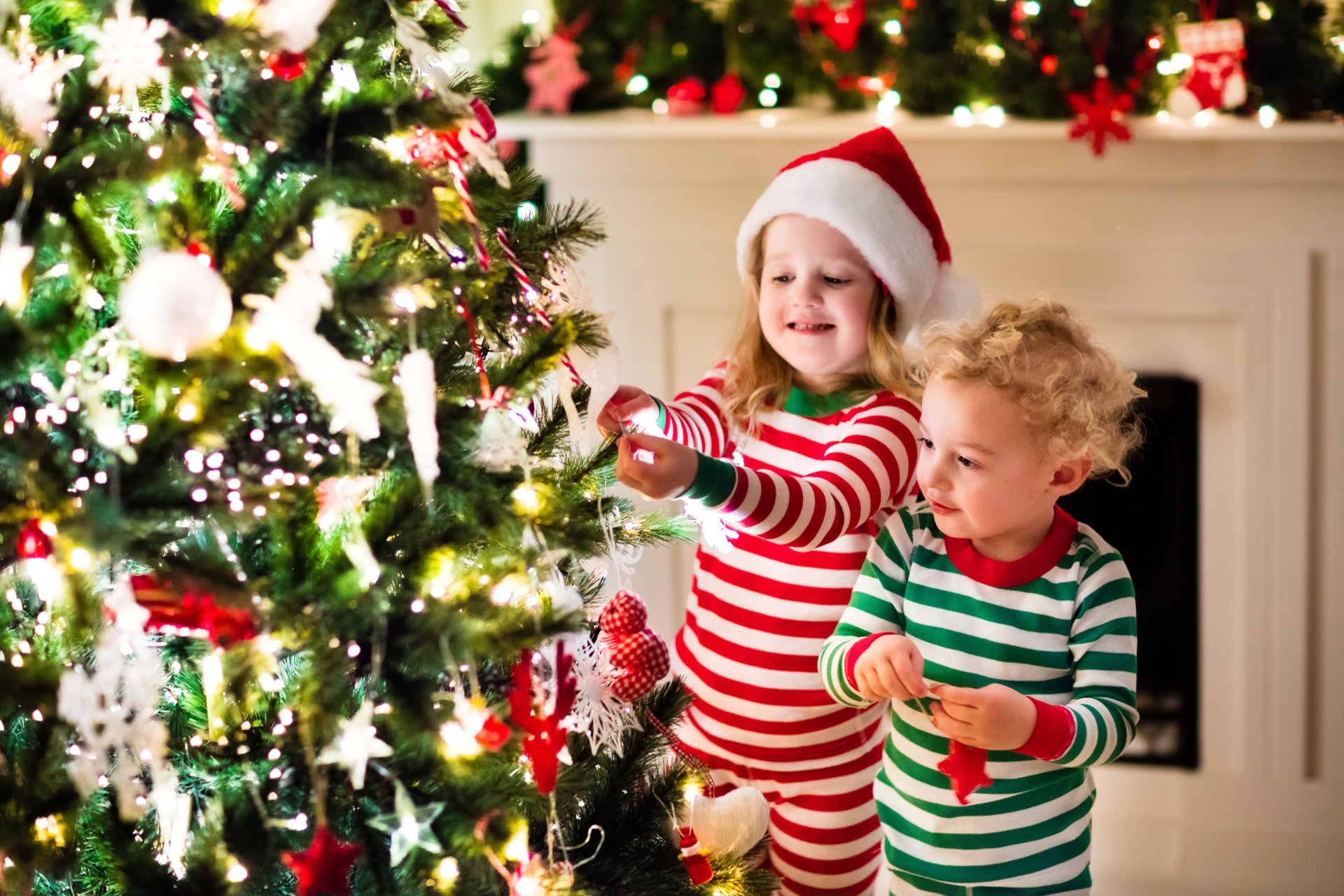 2K Quad HD PC desktop wallpaper: little girl in a Santa hat and little boy in striped pajamas place ornaments on a glowing Christmas tree, joyful children celebrating the holiday.