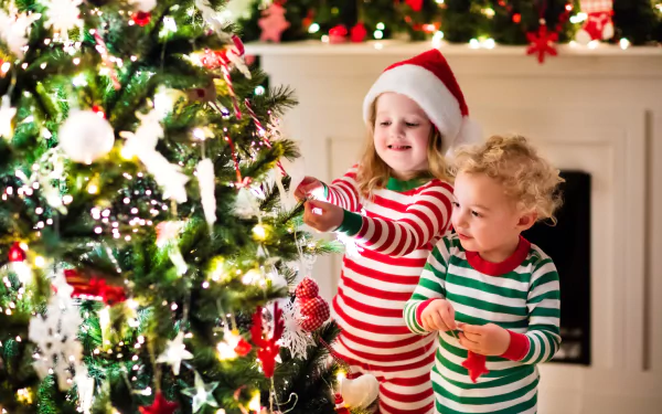 2K Quad HD PC desktop wallpaper: little girl in a Santa hat and little boy in striped pajamas place ornaments on a glowing Christmas tree, joyful children celebrating the holiday.