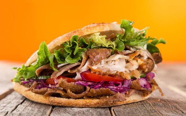 5K Ultra HD PC wallpaper of a doner kebab: sliced meat, bread, lettuce, cabbage and tomato in a pita on a wooden surface against an orange background.