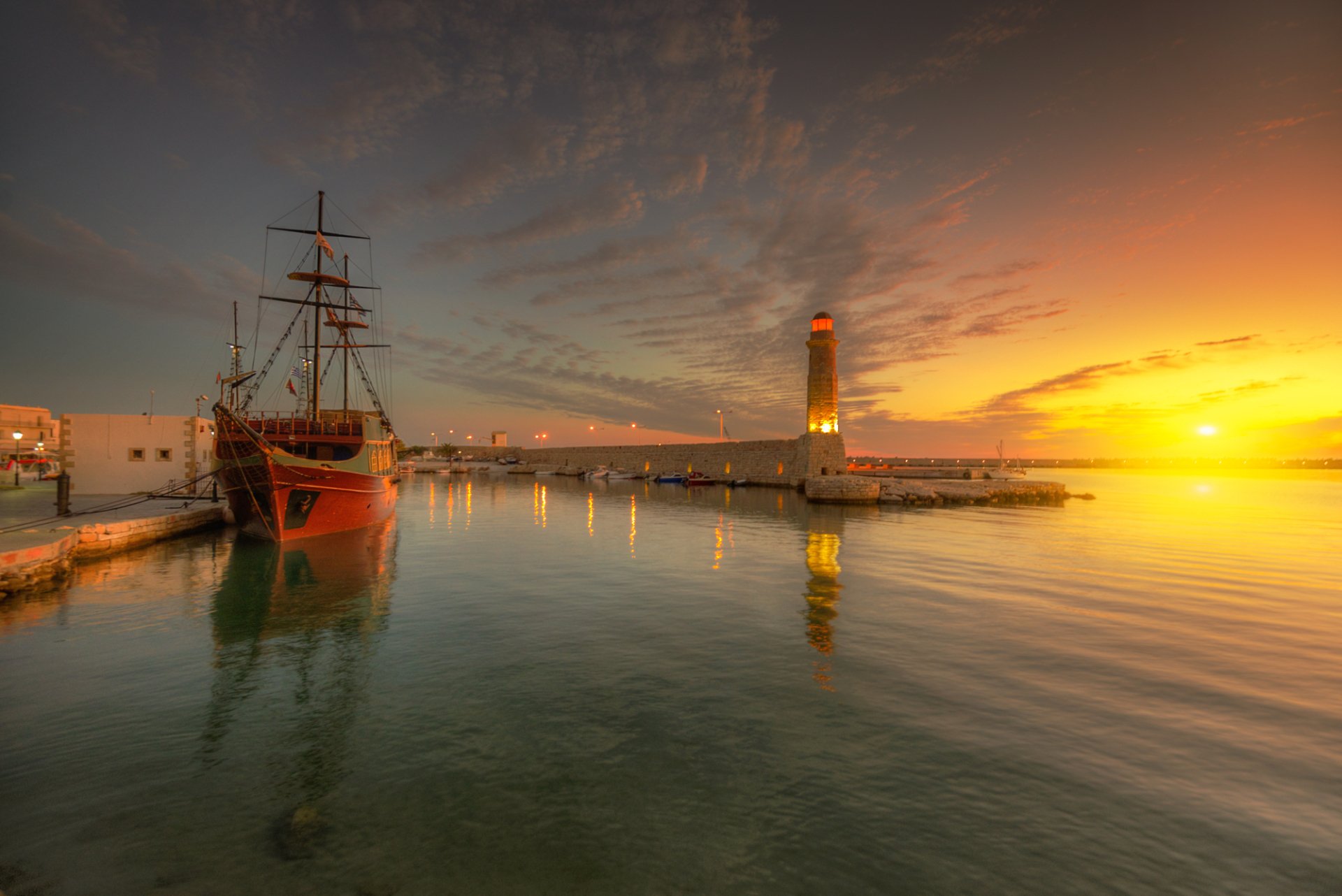 HD desktop wallpaper of a serene ocean sunset featuring a ship docked near a lighthouse, silhouetted against a vibrant sky with orange and yellow hues reflecting on the water.